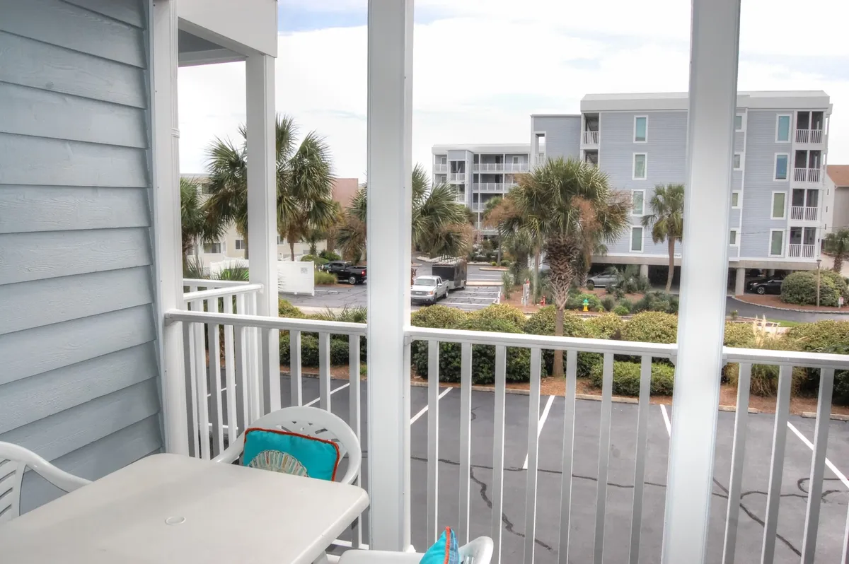 This image shows a screened-in balcony with a table and chairs, framed by a railing, and offers a view of palm trees and a peaceful parking lot below. The seating area is inviting, allowing someone to imagine savoring a morning coffee or relaxing with a glass of wine while enjoying the Myrtle Beach breeze. The overall atmosphere is tranquil and semi-private, with resort greenery visible beyond the railing.