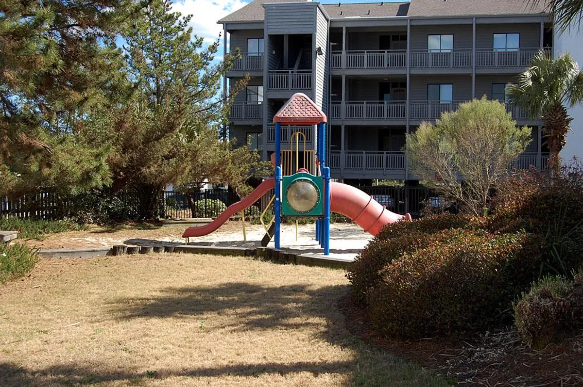 A cheerful outdoor playground surrounded by lush greenery and the property facade in the background. The scene shows children playing and families gathering, highlighting the on-site amenities that add value to a family getaway.