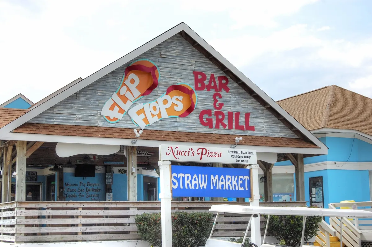 An energetic exterior shot that captures Flip Flops Bar & Grill, Nucci's Pizza, and the Straw Market, along with their outdoor seating areas and vibrant signage. People can be seen dining, socializing, and enjoying the lively ambiance of this local hub.