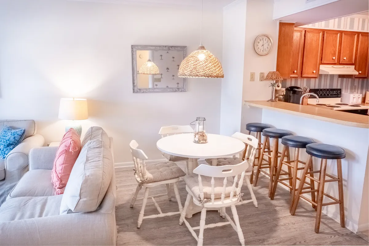 A cozy dining area featuring a round white table surrounded by chairs, illuminated by woven pendant lights. Blue accent pillows add a pop of color, and the kitchen bar area is visible in the background, seamlessly integrating dining and kitchen spaces.