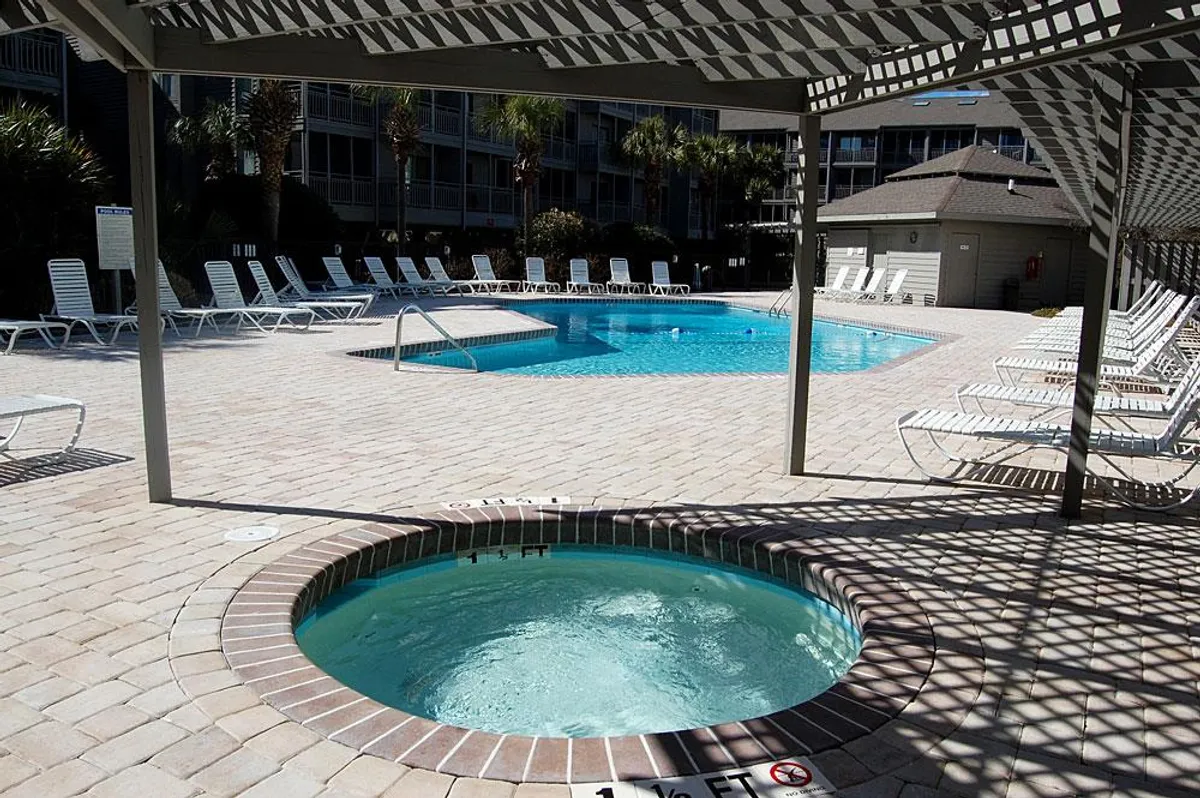 A sunlit outdoor swimming pool and hot tub surrounded by lounge chairs, with palm trees and clear skies in the background. The space looks inviting, perfect for both active fun and restful lounging.
