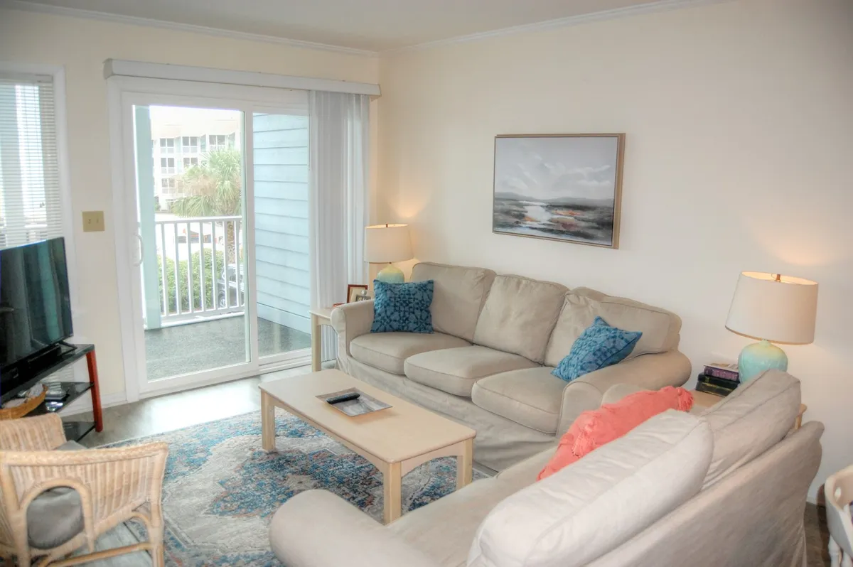 This image features a spacious living room with a plush sofa, coffee table, television, tasteful artwork, and a sliding glass door that allows natural light to flood the space. The overall setup feels inviting and ideal for relaxing, socializing, and enjoying both indoor comforts and outdoor views.
