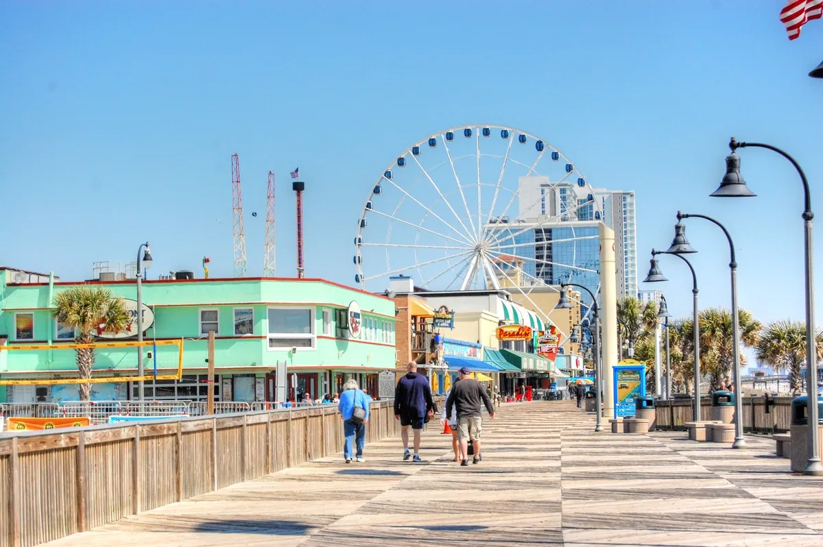 A vibrant scene along the Grand Strand boardwalk, featuring a towering ferris wheel, palm trees, lively crowds, and a mix of local shops and green buildings. The energy and diversity of activities highlight the entertainment and dining options nearby.