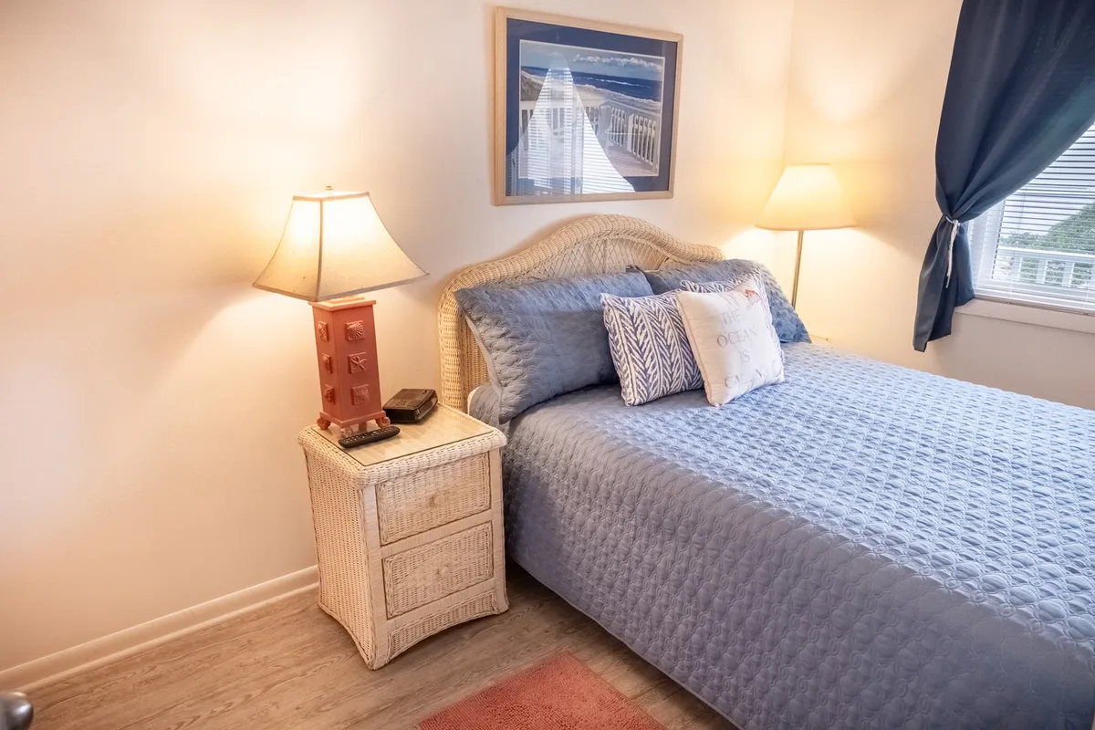 A cozy queen bedroom featuring blue bedding, a nightstand with a lamp, and framed artwork on the wall. Gentle natural light fills the space, enhancing its sense of peace and comfort.