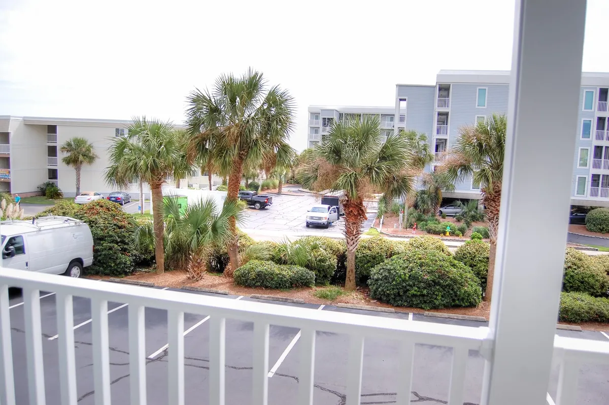 This image features a serene view from a balcony overlooking manicured shrubs, palm trees, and parts of the resort community. The perspective is from the balcony, with the outdoor area and parking lot visible, reinforcing the sense of being nestled within a tranquil, green resort setting. The lighting suggests a calm, inviting time of day, ideal for relaxation.