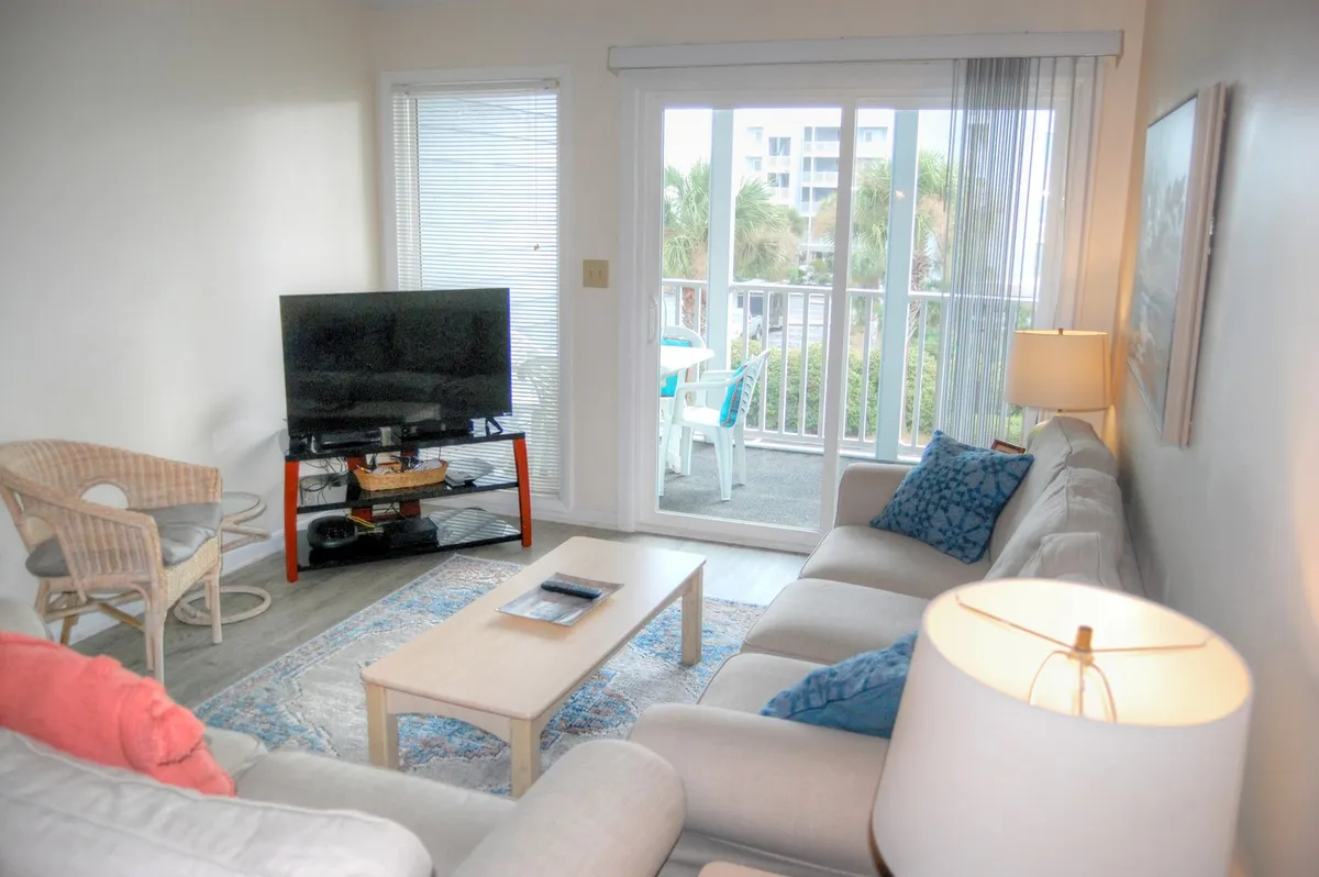 The image showcases a bright, inviting living room with a comfortable sofa and coffee table set up for relaxation or game nights. Sliding glass doors open to a private balcony with views toward the beach, creating a seamless indoor-outdoor atmosphere perfect for unwinding while watching sunsets. A large television adds to the cozy, home-like ambiance, making this space ideal for movie nights with family or friends.