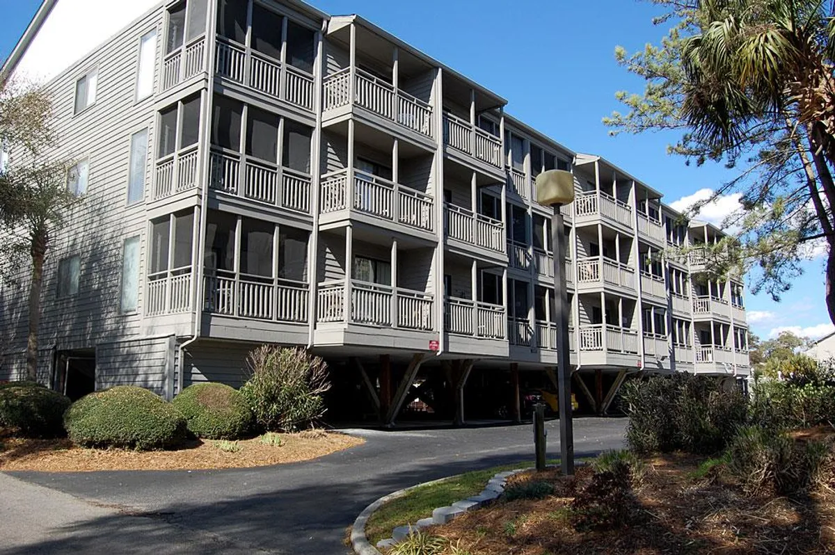 A welcoming exterior view of the condo building, featuring attractive balconies, landscaped shrubs, and a pathway leading to the entrance. The scene hints at private outdoor spaces for relaxation and easy access to the surrounding attractions.