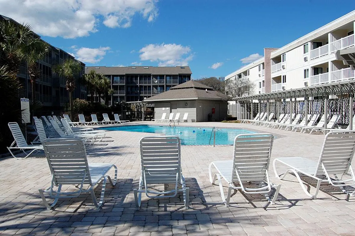 A large, inviting swimming pool is surrounded by an array of lounge chairs and mature palm trees, with the building’s coastal architecture framing the backdrop. The pool area is bathed in natural sunlight, creating a tranquil and resort-like atmosphere ideal for both relaxation and socializing.