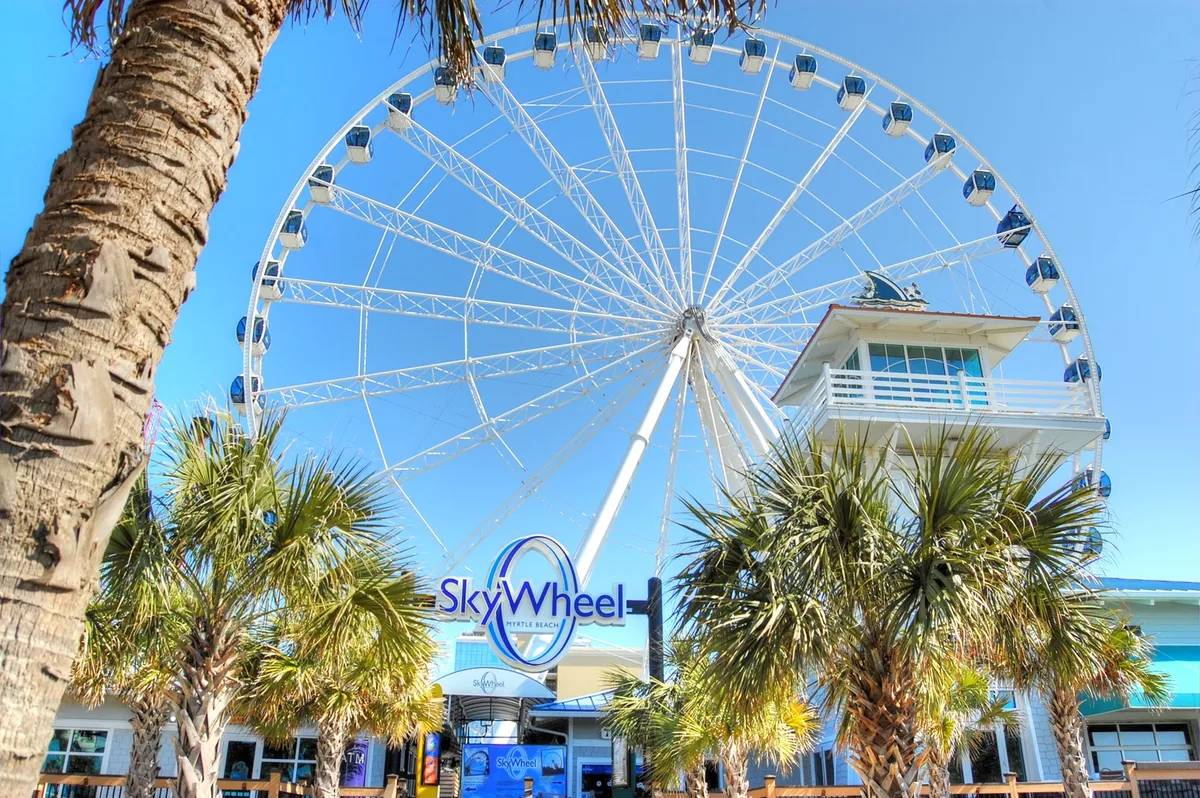 This image captures the iconic SkyWheel, framed by palm trees and the entrance to a local building. The vibrant outdoor setting is filled with anticipation and energy, reflecting the excitement of visiting a well-known Myrtle Beach attraction.