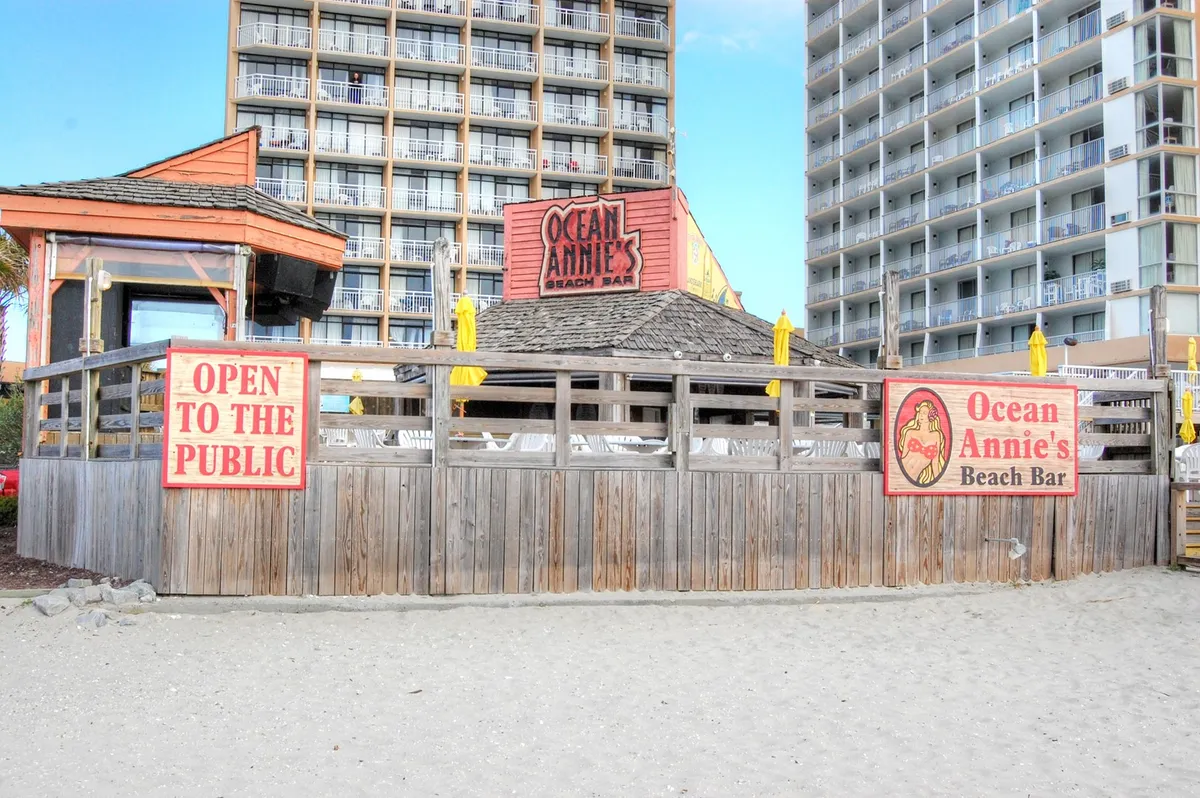 This lively exterior shot features the iconic Ocean Annie's sign and its open-to-the-public beach bar structure, set just steps from the sand. The inviting setting hints at live music, social gatherings, and relaxed beach vibes, with patrons enjoying drinks and the ocean view.