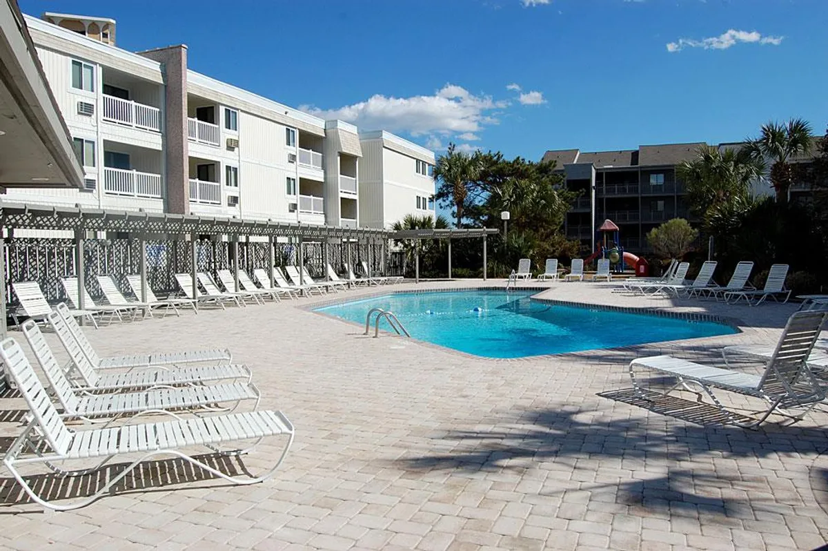 A sparkling swimming pool lined with lounge chairs and bordered by graceful palm trees, with the resort buildings visible in the background. The scene exudes a relaxed, sunlit ambiance perfect for both solo relaxation and social moments.
