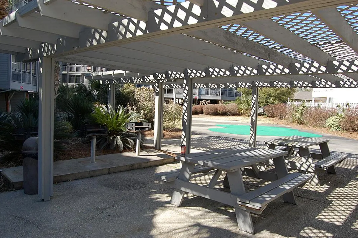 A welcoming outdoor pergola shades picnic tables nestled amid lush greenery, providing a casual and comfortable space for outdoor dining or social gatherings. The setting hints at opportunities for barbecues or evening get-togethers under the open sky.