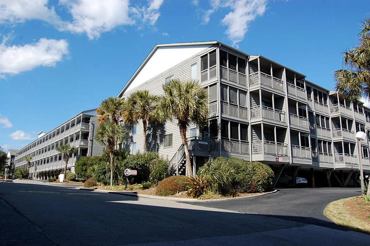 The exterior view of a multi-story resort building framed by swaying palm trees and manicured gardens. Balconies overlook the grounds, hinting at private spaces that connect guests directly to the outdoor atmosphere of the coast.