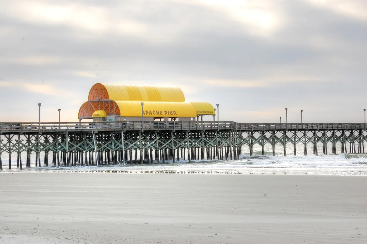 A wide view of the iconic Apache Pier stretching out over the ocean, featuring its distinctive yellow roof and sturdy pier supports. The scene captures the tranquil water, open sky, and inviting atmosphere of the area, evoking a sense of relaxation and coastal community.